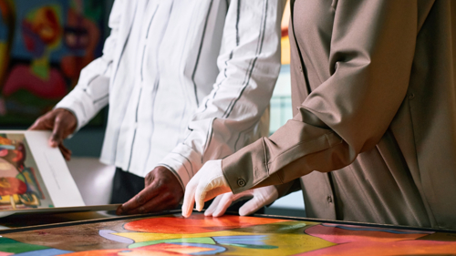 A close up of two people, one browsing through a book, the other in white gloves, hands resting on a painting 