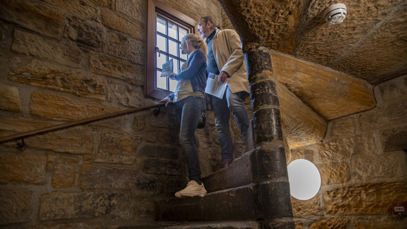 Two visitors climb a narrow stone staircase inside a historic building, pausing by a small window set into thick stone walls. The staircase curves upward with a wooden handrail, and warm lighting highlights the textured masonry and enclosed space.