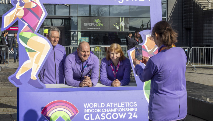 Three volunteers standing behind a purple selfie frame outside the Emirates Arena as another volunteer takes their photo during the 2024 World Athletics Indoor Championships.
