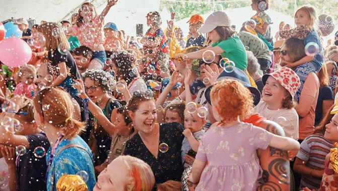 A joyous crowd of children and adults celebrate amidst a sea of bubbles and disco balls at a festival.