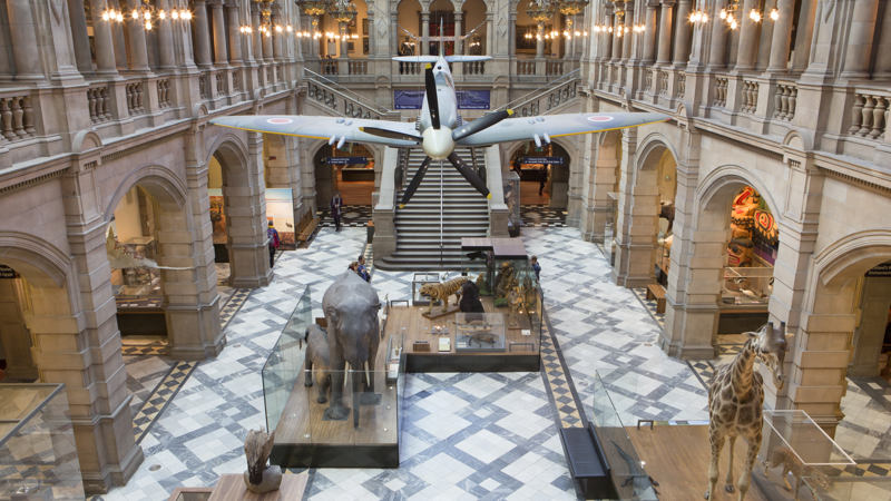 A view of one hall in Kelvingrove Art Gallery and Museum. There are several stuffed animals, including an elephant and a giraffe. There is also a model of a war plane, hanging from the roof. 