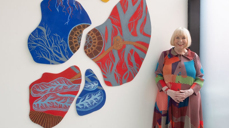 A woman stands beside a wall installation made of several irregular, organic‑shaped panels covered in intricate, vein‑ and branch‑like patterns. Each panel is painted in vivid colors—reds, blues, yellows, and greys—with fine contrasting line work. Her outfit echoes the artwork’s palette and patterns, creating a visual harmony between her clothing and the installation. The setting appears to be a bright, modern gallery space with large windows letting in natural light.