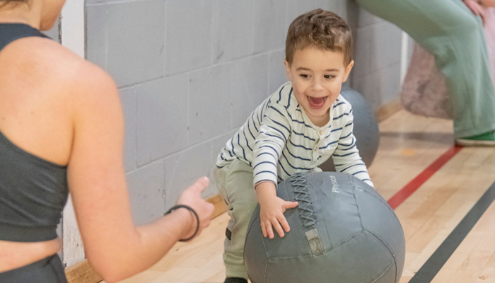 A toddler rolls a large exercise ball across a gym floor toward a seated adult, while other parents and children sit and watch in the background.