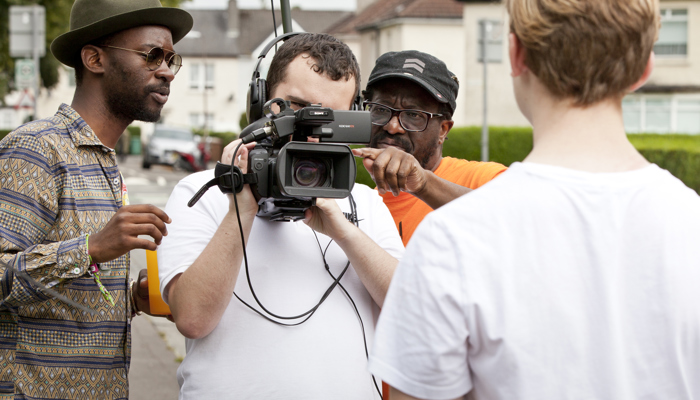 Two adults give advice to a young person with a film camera, who is filming another young person. 