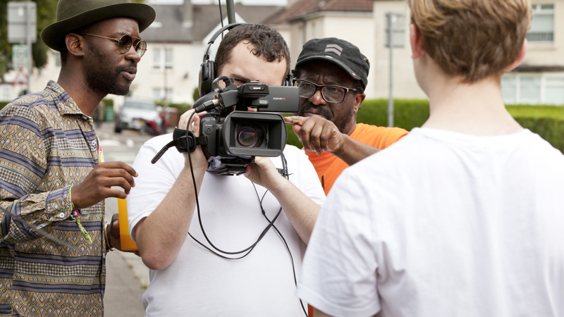 Two adults give advice to a young person with a film camera, who is filming another young person. 