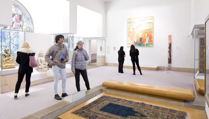 Visitors in St Mungo Museum looking at a tapestry carpet on the floor in display case. The museum is bright and white and several other objects are on display in the background
