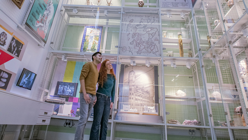 Two visitors stand in a bright, modern museum gallery with high ceilings and a large skylight overhead, looking up at tall glass display cases filled with diverse objects, including framed posters, maps, natural specimens, and decorative items arranged across multiple levels.