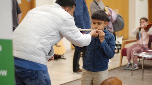 An adult and a young person are trying out musical instruments in a library. The young person is holding up a flute