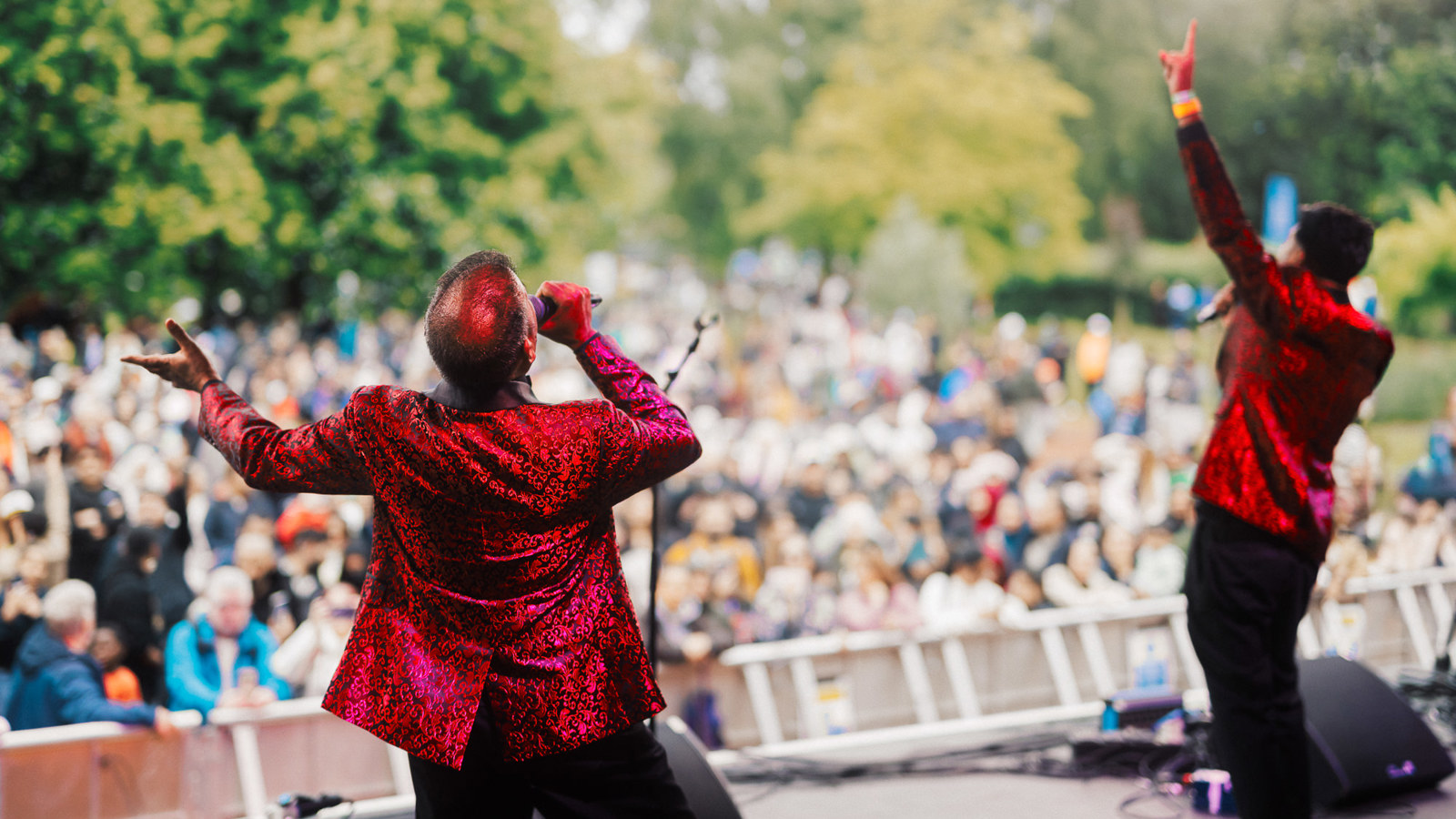 Performers wearing red jackets sing in front of a large crowd at an outdoor music event