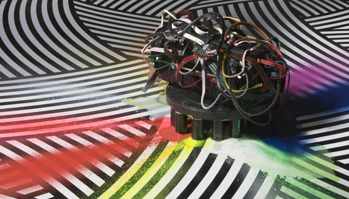 Tangled coloured leather straps are attached to a black holdall bag to create a compact sculptural bundle that  sits on a low circular base. The base stands are made of spray cans which have sprayed red, yellow, green, and blue paint in bursts onto a black-and-white patterned floor.
