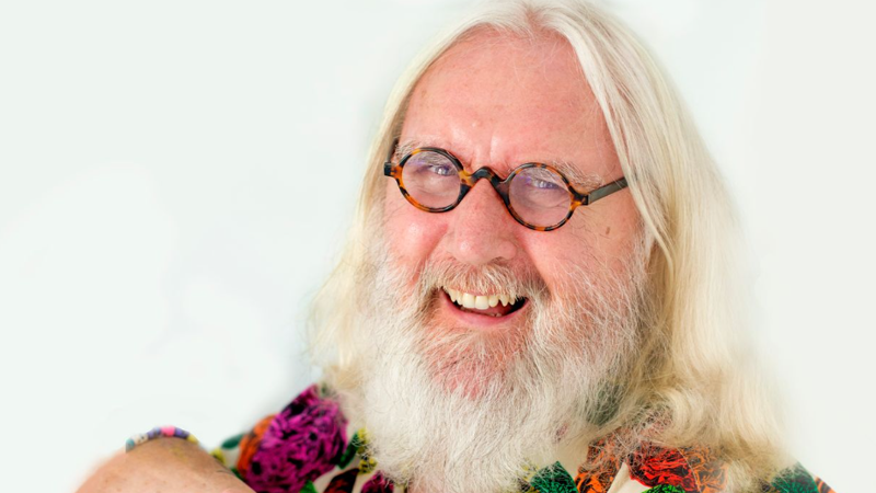 close up image of a man, Billy Connolly, with round spectacles, long white hair and a white beard wearing a colourful shirt, laughing. 