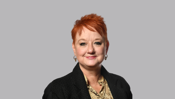 Studio headshot of a person with short red hair, facing forward against a plain light grey background, wearing a black textured jacket over a patterned shirt and small hoop earrings.