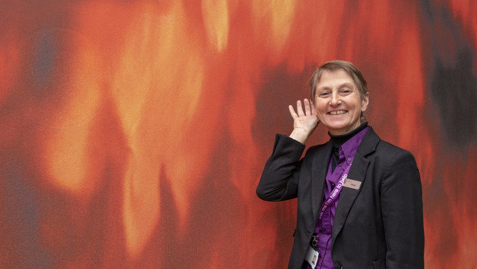 A museum staff member wearing a purple shirt, dark blazer, and lanyard with a name badge raises a hand to an ear in a listening gesture, standing in front of an abstract red and orange wall.