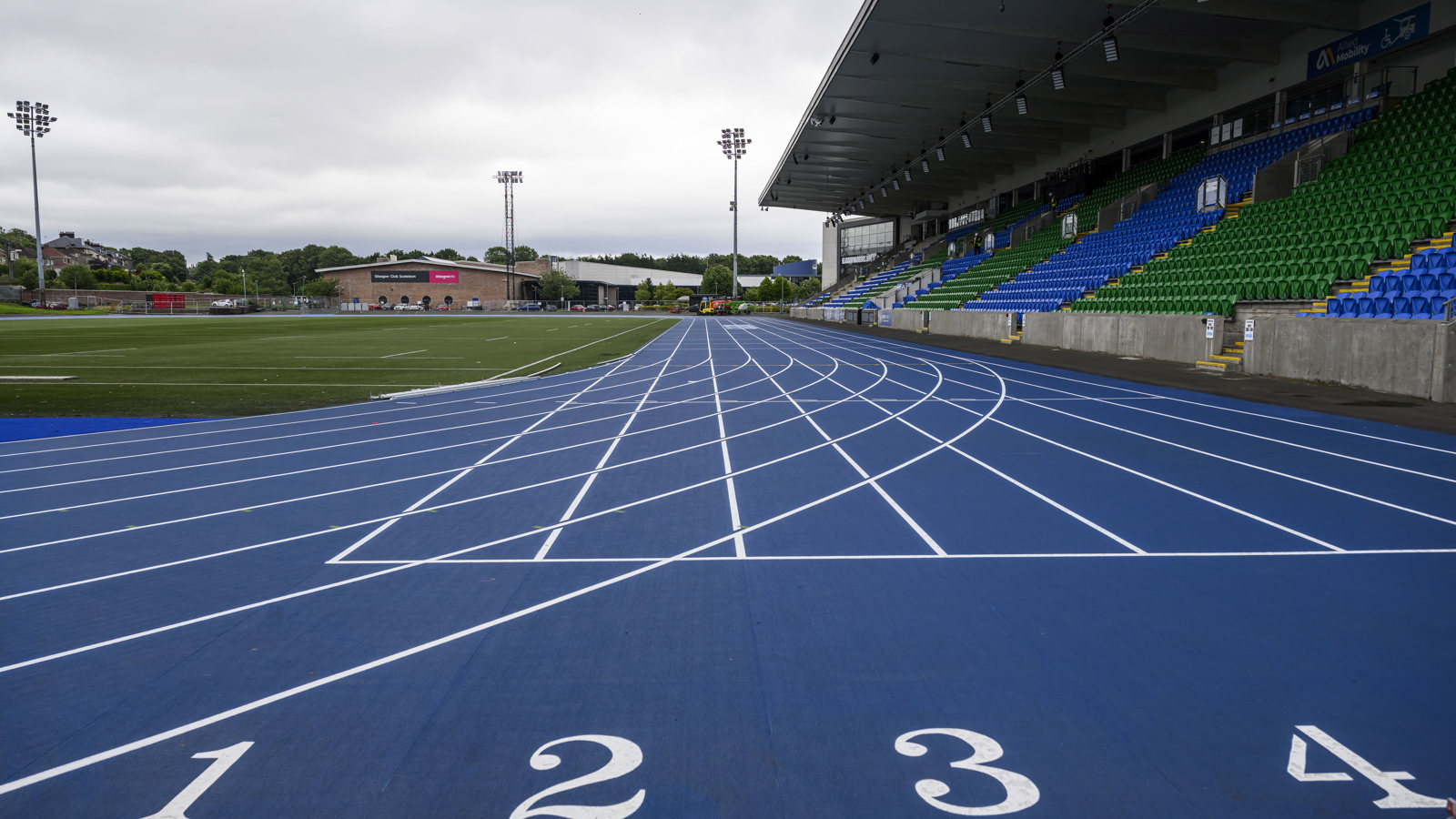 A blue and white running track separates a rugby pitch and a stand