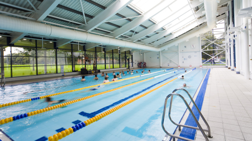 A picture of a swimming pool with swimmers in lanes and a class in the background. 