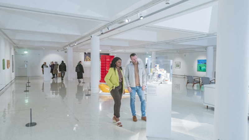 Visitors walk through a bright, modern art gallery with white walls and floors, viewing sculptures and artworks displayed on plinths. Two visitors stand near a central display case examining objects, while other visitors are spaced out across the open exhibition space in the background.