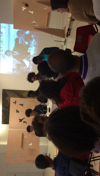 A gallery talk in a white-walled space, with an audience seated in rows facing a panel of speakers at a table, with artworks and a projected image behind them.