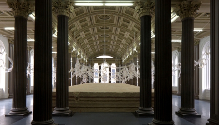 A grand, symmetrical interior space resembling a neoclassical gallery or hall, with a high, arched ceiling richly decorated with ornate panels and moulding. A series of tall, dark columns line both sides, forming a central aisle that leads toward the back of the room. In the middle of the space sits a large, low, rectangular sculpture of types of sawdust creating a layered facade. Above the sawdust, intricate cellophane sculptural forms stretch across the background, contrasting with the darker columns. 