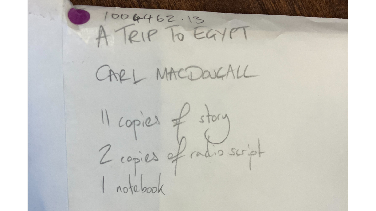A handwritten note pinned with a purple tack lists materials for “A Trip to Egypt” by Carl MacDougall, including 11 copies of the story, 2 copies of the radio script, and 1 notebook