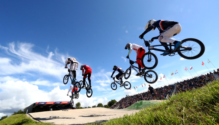 5 people on bikes going over jumps at at the Glasgow BMX Centre. They are outdoors and are wearing safety helmets.