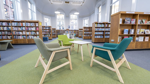Internal photo of a library with large wooden bookshelves filled with colourful books, large windows and cosy green toned armchairs surrounding a coffee table on a green floor runner.