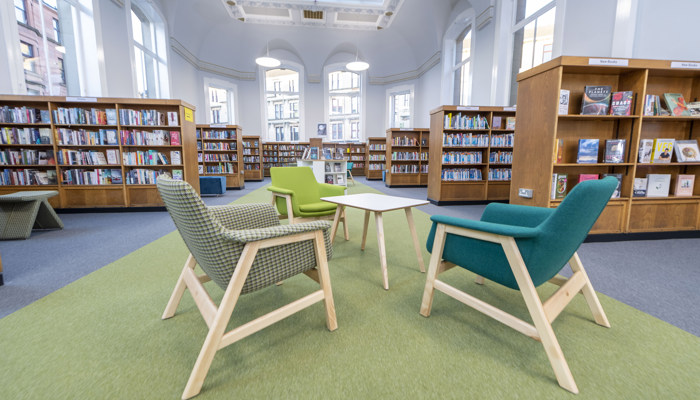Internal photo of a library with large wooden bookshelves filled with colourful books, large windows and cosy green toned armchairs surrounding a coffee table on a green floor runner.