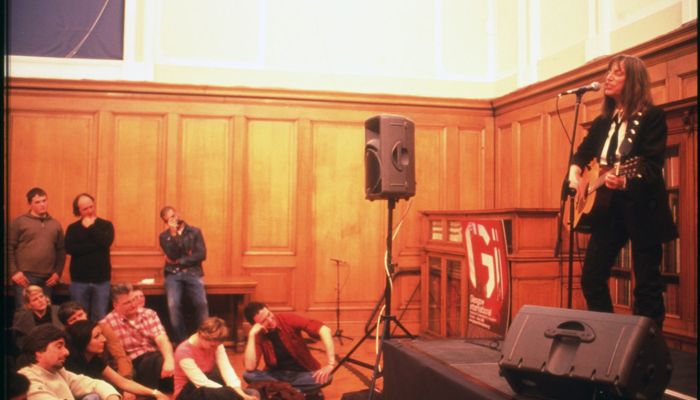 A person with long hair stands on a small stage playing guitar and singing into a microphone, while an audience sits closely on the floor and benches in a wood-panelled room.