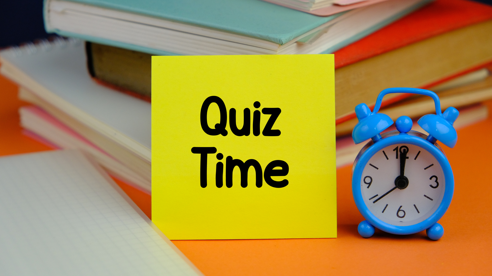 A post-it note with quiz time on top of books with a small blue clock beside it. 
