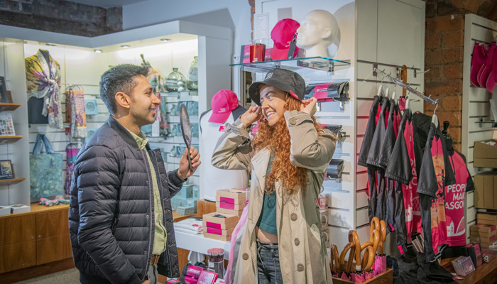 Two people in the Kelvingrove Museum shop. A person with long ginger hair is smiling trying on a hat, whilst another looks on smiling.