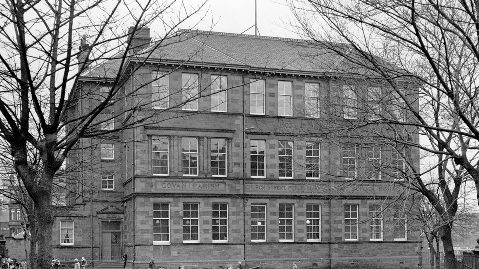 Black and white photo of Govan Parish Church Street Public School with bare tree branches coming in from the sides and children playing in the playground.
