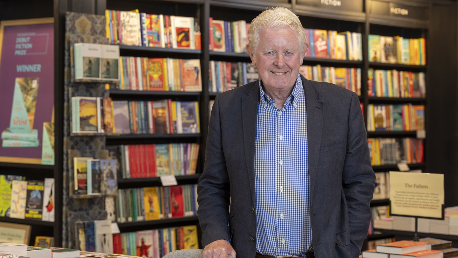 A man with light blonde hair wearing a blue shirt and dark jacket leans on a chair in a library