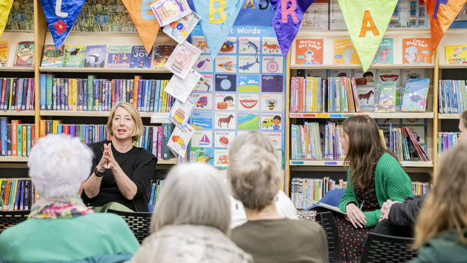 A person is sitting in front of shelves of books in a library and facing a group of people speaking to them 