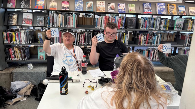Three people are sitting at a table within a library. Two are holding their mugs up towards the camera. 