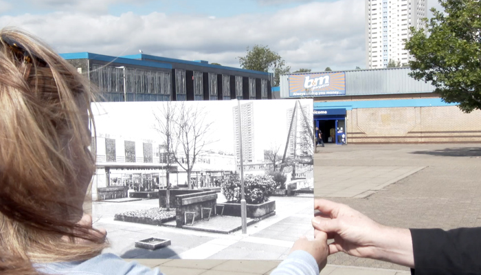 Two people look at an old photograph of the site they are looking at. The site in the past has buildings which are now demolished. 