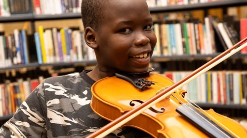 A young person smiling and holding a violin under their chin in a library