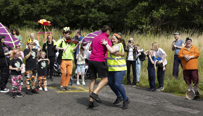 A multi-generational crowd look on as two people dance together, accompanied by a violinist 