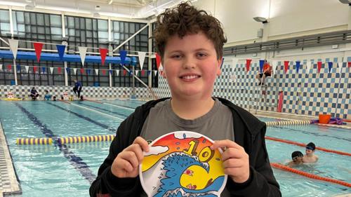 A young person standing next to an indoor swimming pool smiling and holding a colourful swimming cap in front of them