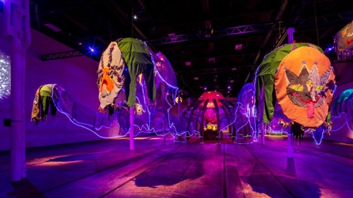 A vast gallery space is bathed in ultraviolet light, filled with a huge sculpture which resembles a jelly fish. In the foreground two colourful embroidered panels hang at the end of two tentacles