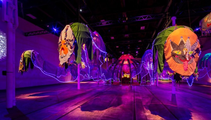 A vast gallery space is bathed in ultraviolet light, filled with a huge sculpture which resembles a jelly fish. In the foreground two colourful embroidered panels hang at the end of two tentacles