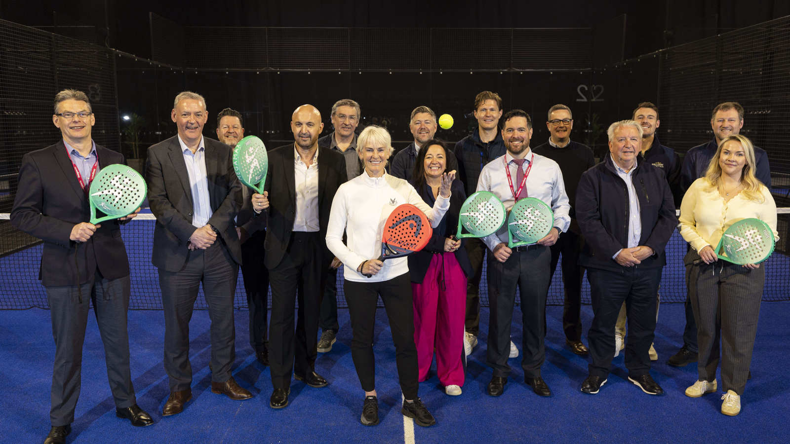  A large group of people gathered for the opening of a new indoor padel court. Some of them are holding padel raquets and the person in the centre is throwing a yellow ball in the air