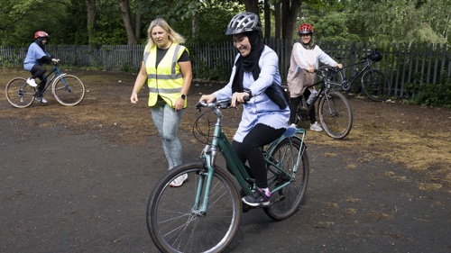 A group of three people taking part in a cycling lesson with an instructor on a tarmac area in a park