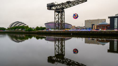 A large circular artwork hangs from a crane above a river and beside two entertainment venues
