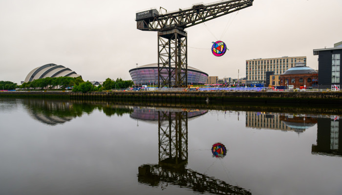 A large circular artwork hangs from a crane above a river and beside two entertainment venues