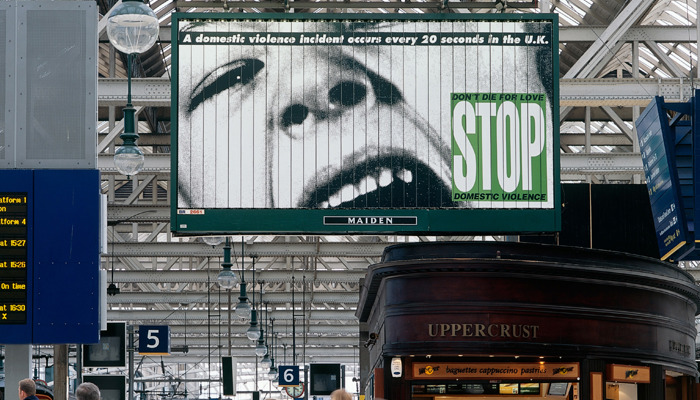 A busy train station interior with people walking past shops, beneath a large billboard showing a black-and-white face and bold text in a green box reading “STOP domestic violence.”