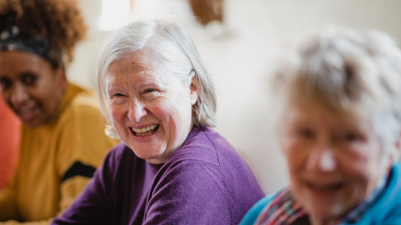 Three people are sitting at a table. The camera is focussed on one person smiling, the background is blurred. 