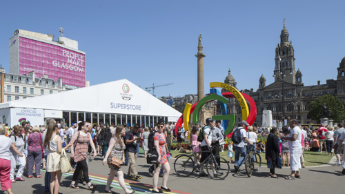 George Square on a sunny day during the 2014 Commonwealth Games, lots of people are walking around next to the Big G sculpture and the People Make Glasgow Met Tower overlooks the square.