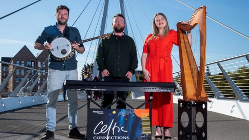 Three people standing on a metal pedestrian bridge on a clear day. One of them is holding a banjo, one is standing behind a keyboard, one has a large harp.