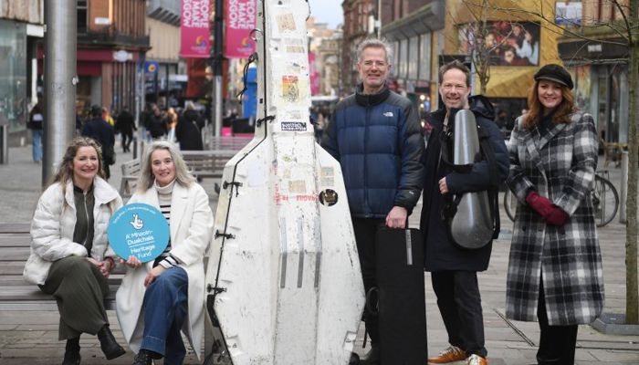Five people pose on a city street around a large white instrument case, with two holding cello cases and pedestrians in the background.