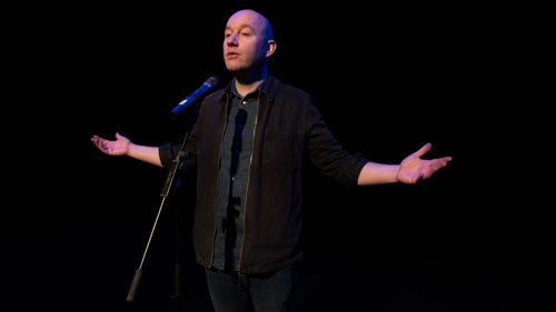 A performer wearing dark clothing stands in front of a microphone with their arms outstretched