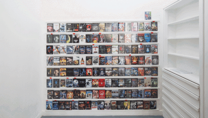 A minimalist white gallery-like room with a smooth grey floor. Centered on the back wall is a large, rectangular display composed of many DVD or Blu-ray cases arranged tightly in a grid of rows and columns. The cases vary in color and design, creating a dense mosaic of film cover artwork. To the right, a built-in white shelving unit with several horizontal shelves and drawers sits flush against the wall. 